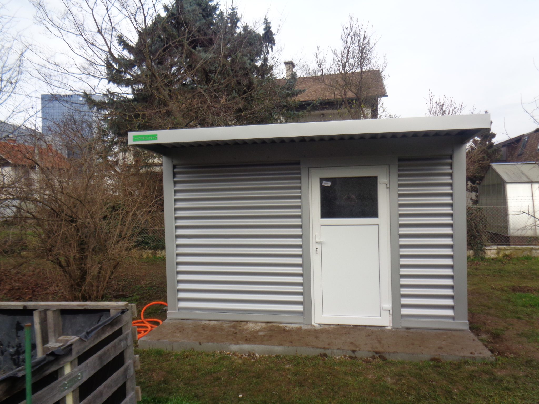 Srebrne kovinske Hosekra vrtne lope z belimi vrati, postavljena na obrobje parcele.