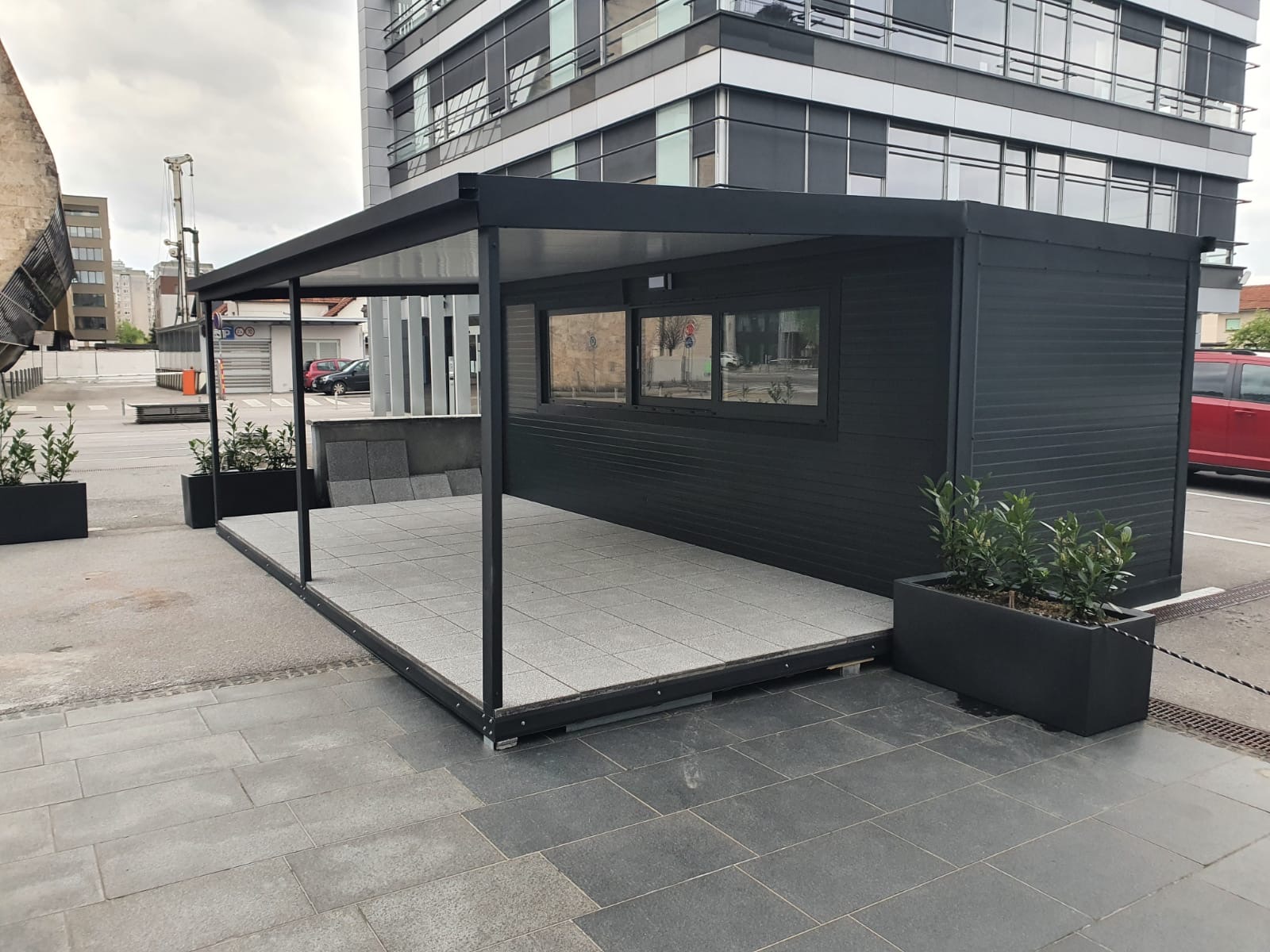 Gostinjski Kontejner za prodajo v antracit sivi barvi, postavljen v urbanem okolju na ulici, med nebotičniki.